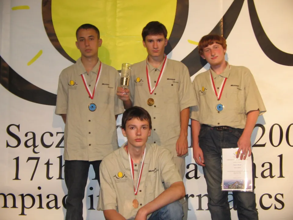 Four members of Ukraine’s team in matching light shirts with their medals in front of the IOI 2005 banner in Nowy Sącz; one holds a cup, another a certificate